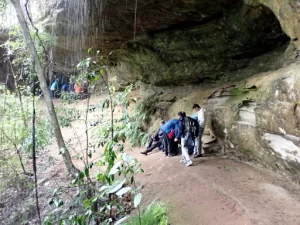 A group of Hikers are standing under a rock ledge with water pouring down while exploring country during a Gold Bushwalking Award Journey