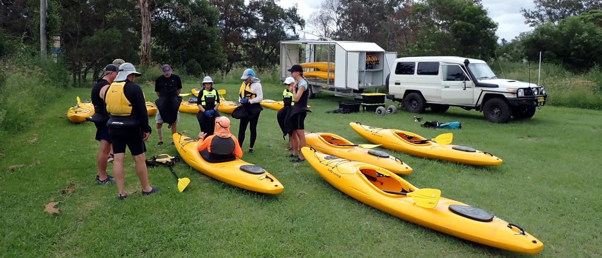 A group pf people are standing around an instructor learning how to kayak