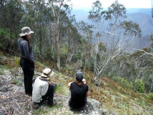 three hikers are sitting on top of a mountain looking down at the snowy river on the valley floor during the Mount Kosciuszko to Bass Strait hike