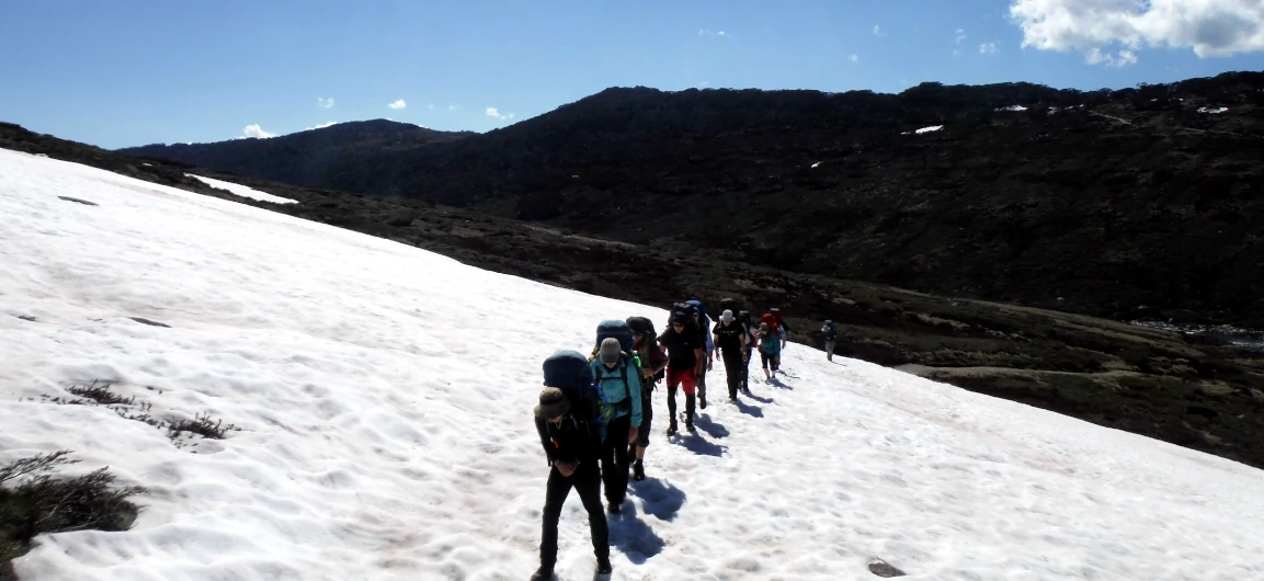 A group of hikers are walking up a steep hill in snow on the Mount Kosciuszko to Bass Strait