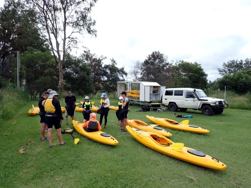 A group of people are standing around an instructor learning about the kayaks that are on the ground in front of him during the Barrington River Kayak Adventure